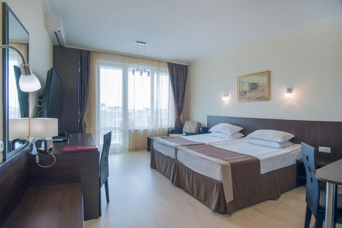Habitación de hotel con dos camas, un escritorio con lámpara, una silla y un gran ventanal con cortinas.