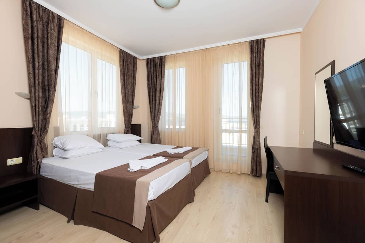 Habitación de hotel con dos camas, un escritorio, un televisor y ventanas amplias con cortinas.