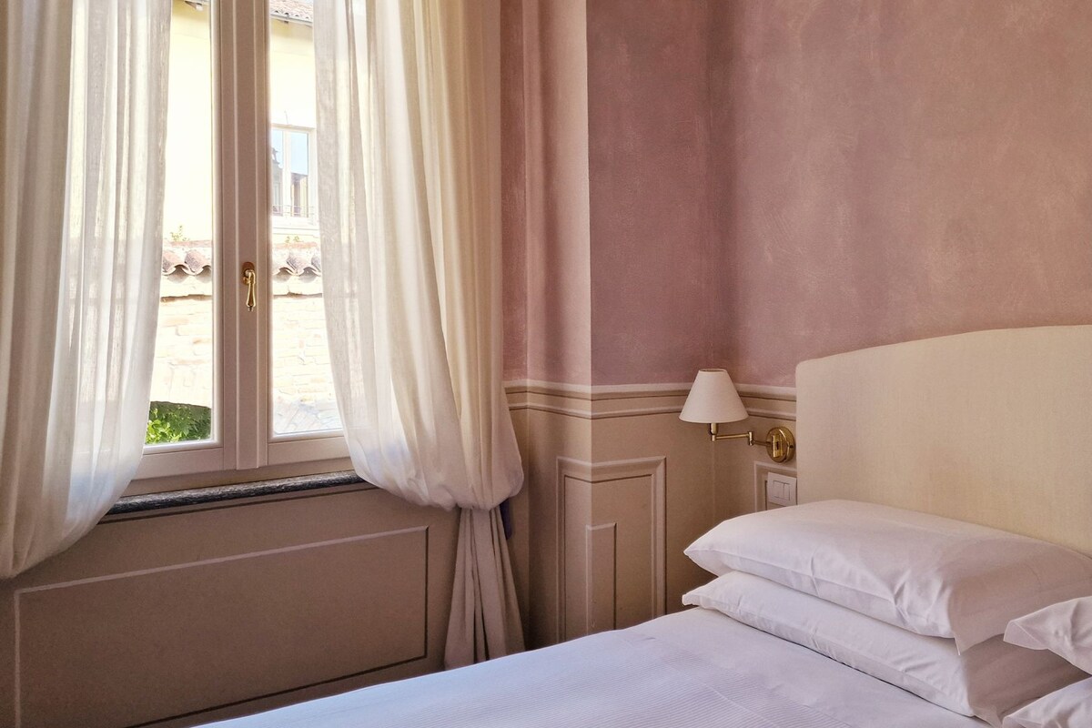 A hotel room with a bed, white curtains, a window, and a wall-mounted lamp.