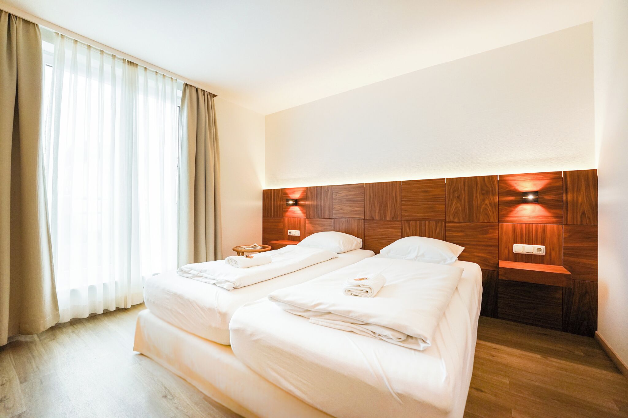 A hotel room with two beds, a wooden headboard, bedside lamps, and a large window with curtains.