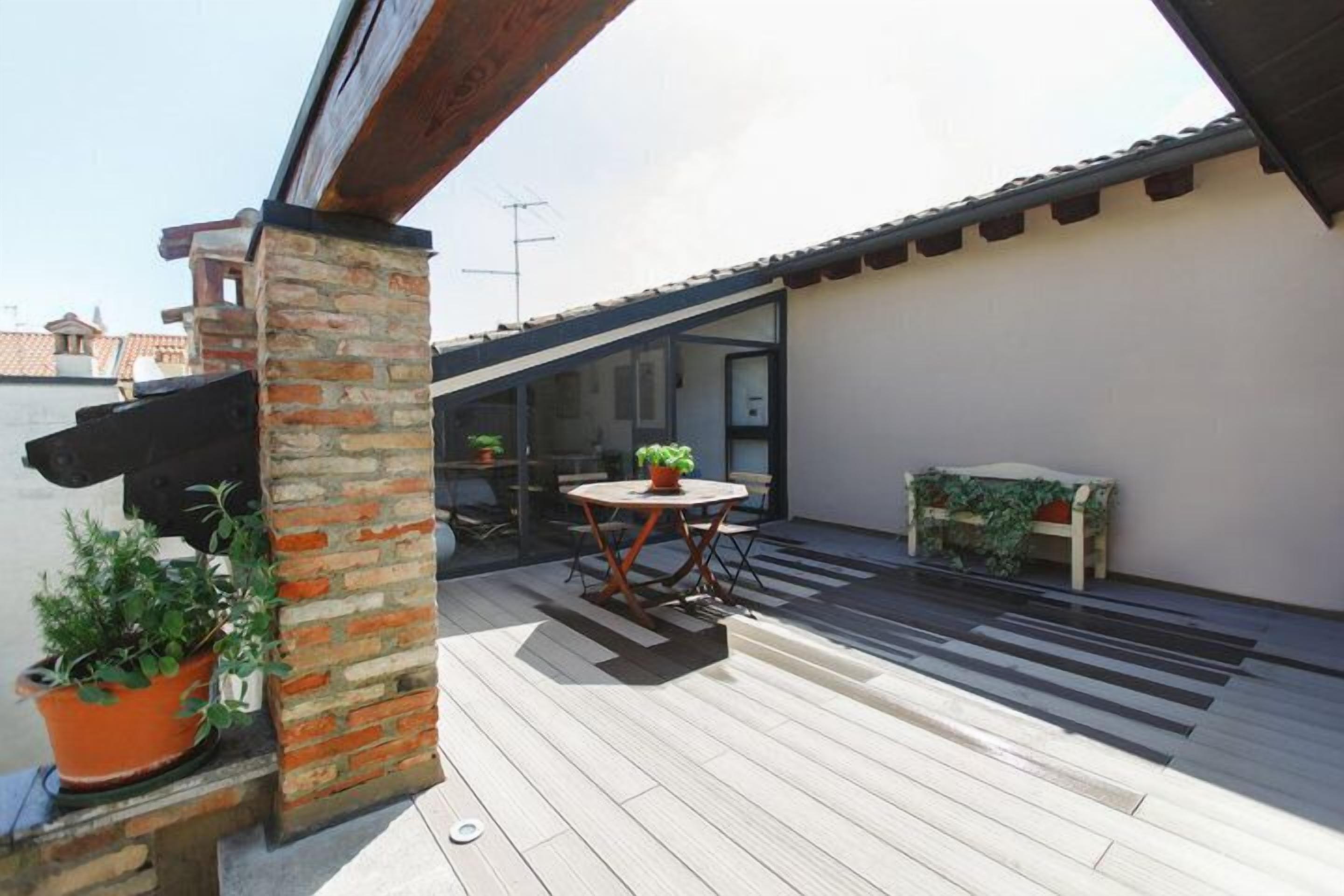 Una terraza en la azotea con cubierta de madera, un pilar de ladrillo, una mesa redonda con sillas y plantas en macetas.