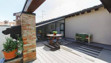Una terraza en la azotea con cubierta de madera, un pilar de ladrillo, una mesa redonda con sillas y plantas en macetas.