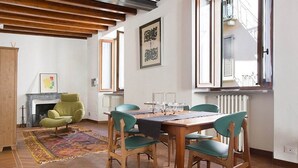 A room with a wooden ceiling, a fireplace, a dining table with chairs, and a window with a view of the outside. - B&B San Clemente (Crema)