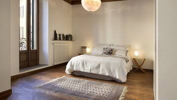 A bedroom with a wooden floor, a white bed with patterned bedding, a hanging light fixture, and a radiator.