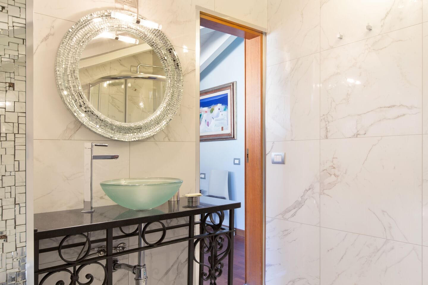 A bathroom with a glass bowl sink, a mosaic mirror, and a marble floor.