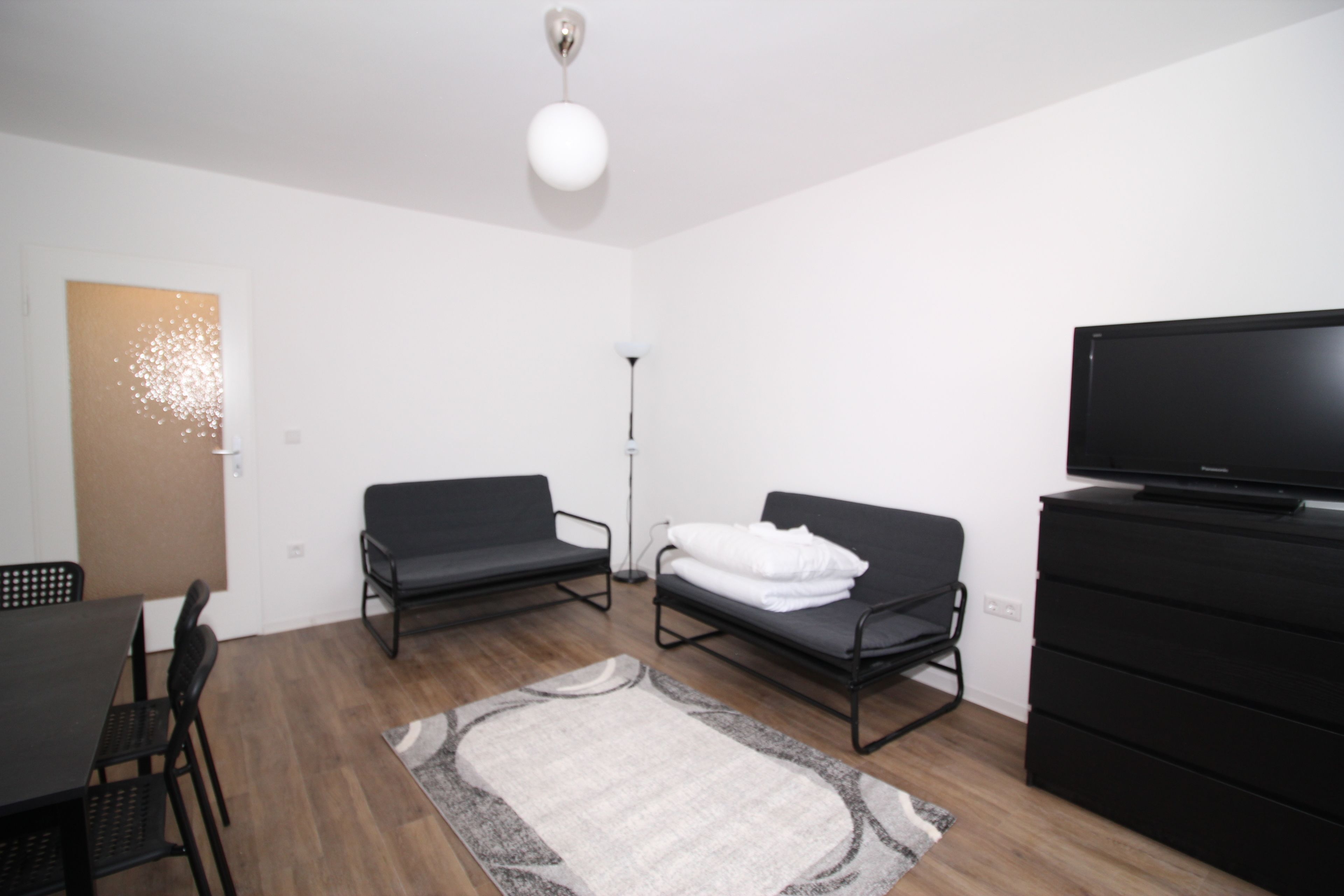 A modern, minimalistic room with a flat-screen TV, two black sofas, a dining table with chairs, and a large window.