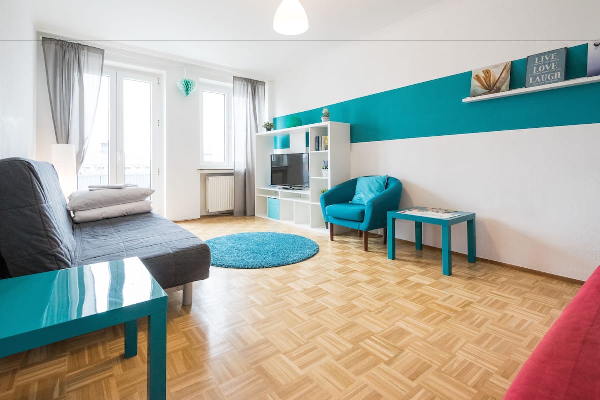 A modern living room with a sofa, a teal armchair, a small table, and a bookshelf.