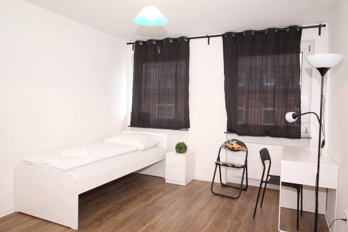 A small, neatly arranged bedroom with a single bed, a desk, a chair, and a lamp.