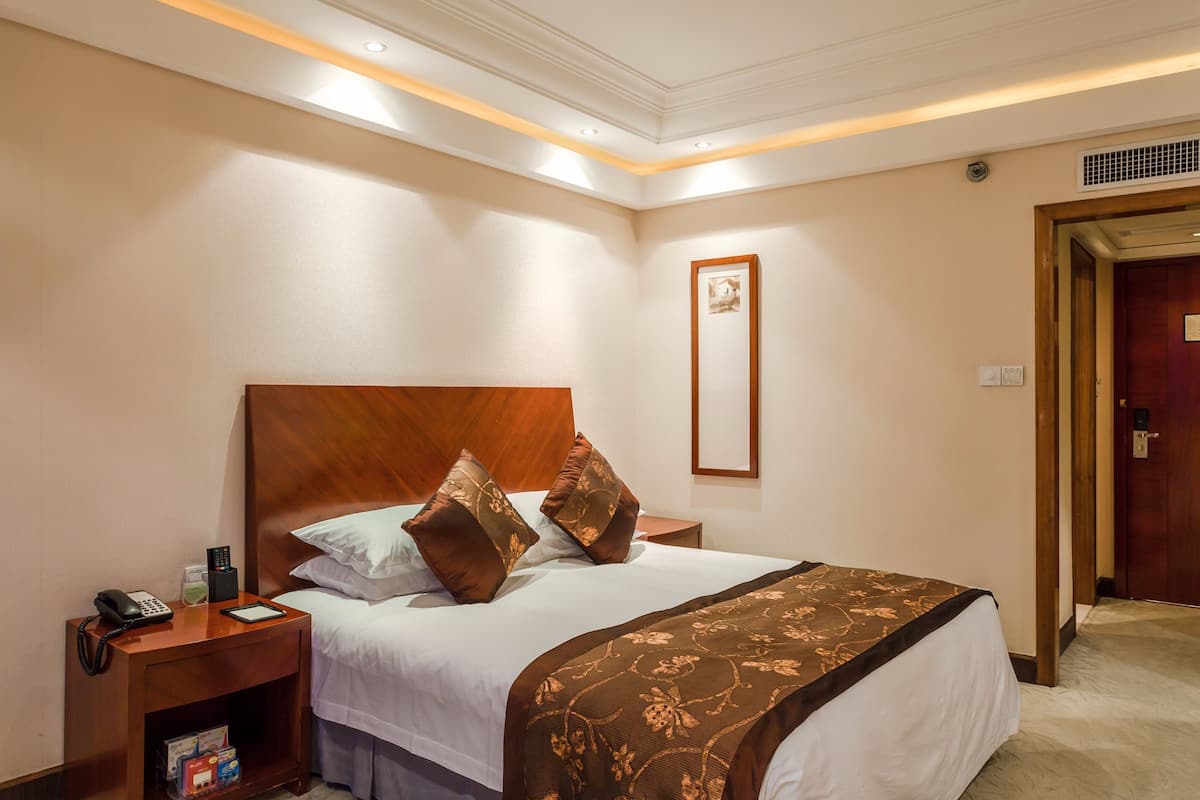 Ein Hotelzimmer mit einem großen Bett, einem Holzkopfteil, Nachttischen mit Telefon und Fernbedienung sowie einem gerahmten Bild an der Wand.