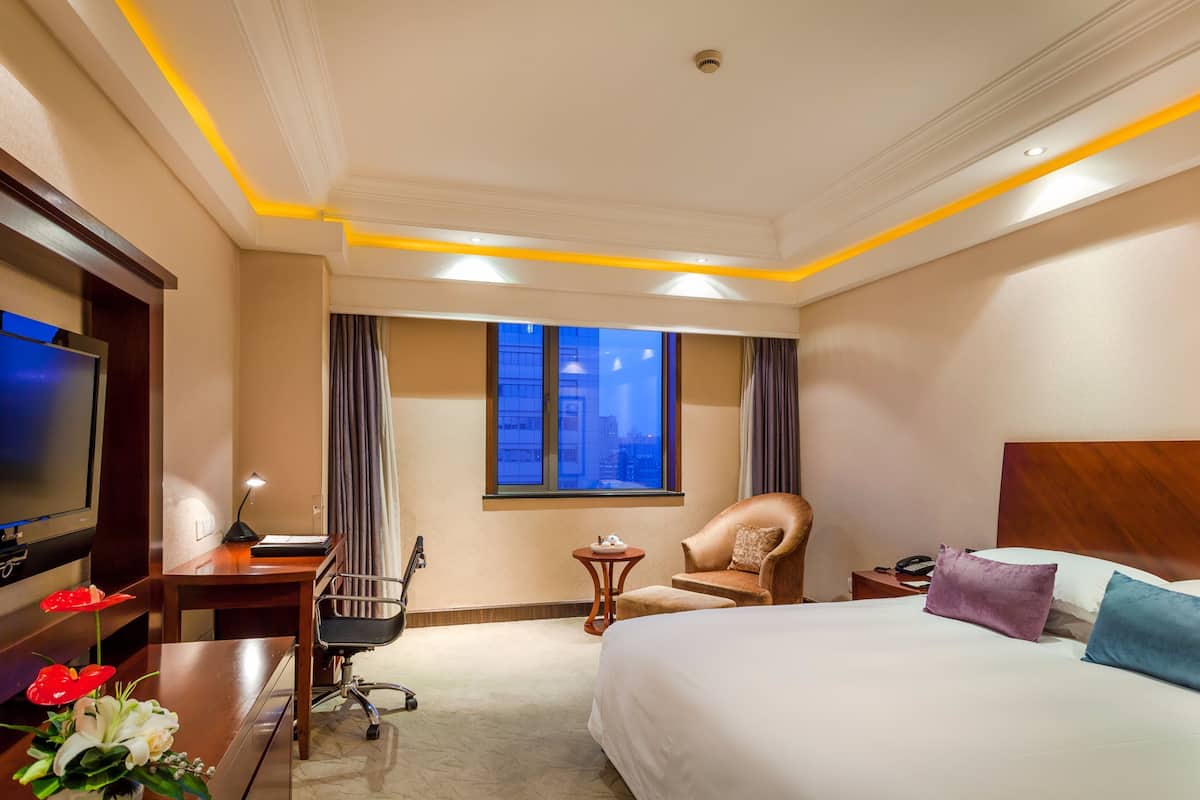 Ein Hotelzimmer mit einem Bett, einem Schreibtisch, einem Sessel, einem Fernseher, einem Fenster mit Vorhängen und einem Nachttisch mit Lampe.