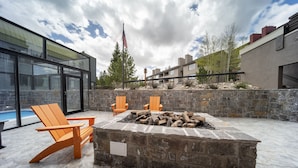 An outdoor patio with a stone fire pit, two wooden Adirondack chairs, and a swimming pool in the background.