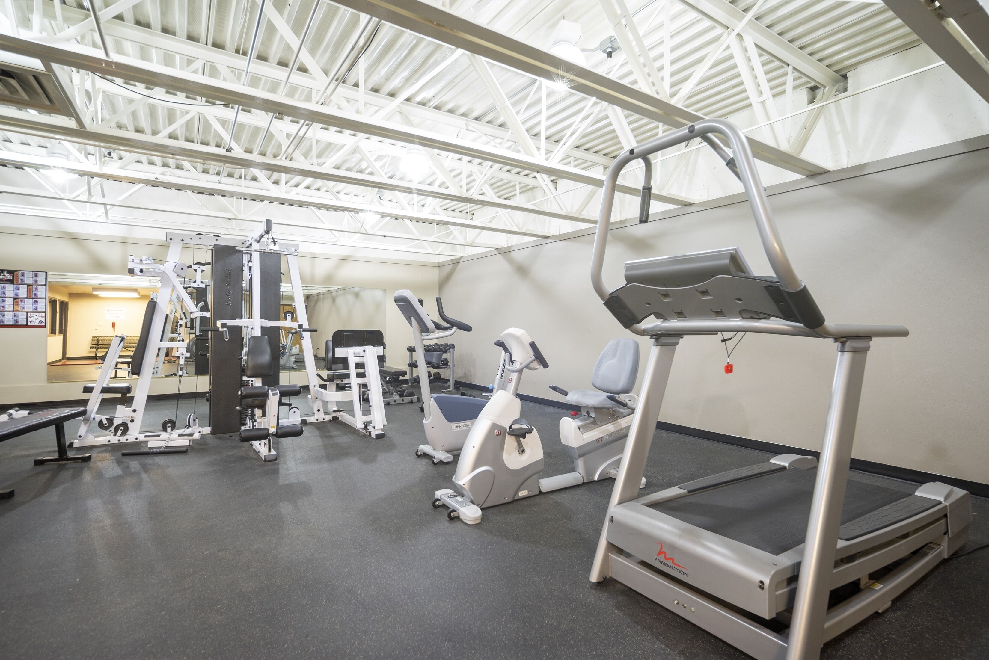 Un gimnasio con diverso equipamiento de ejercicio, incluyendo cintas de correr y máquinas de pesas, bajo un techo alto con vigas metálicas.