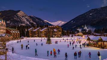 A hotel with multiple buildings, a large open ice skating area, and snow-covered mountains in the background.