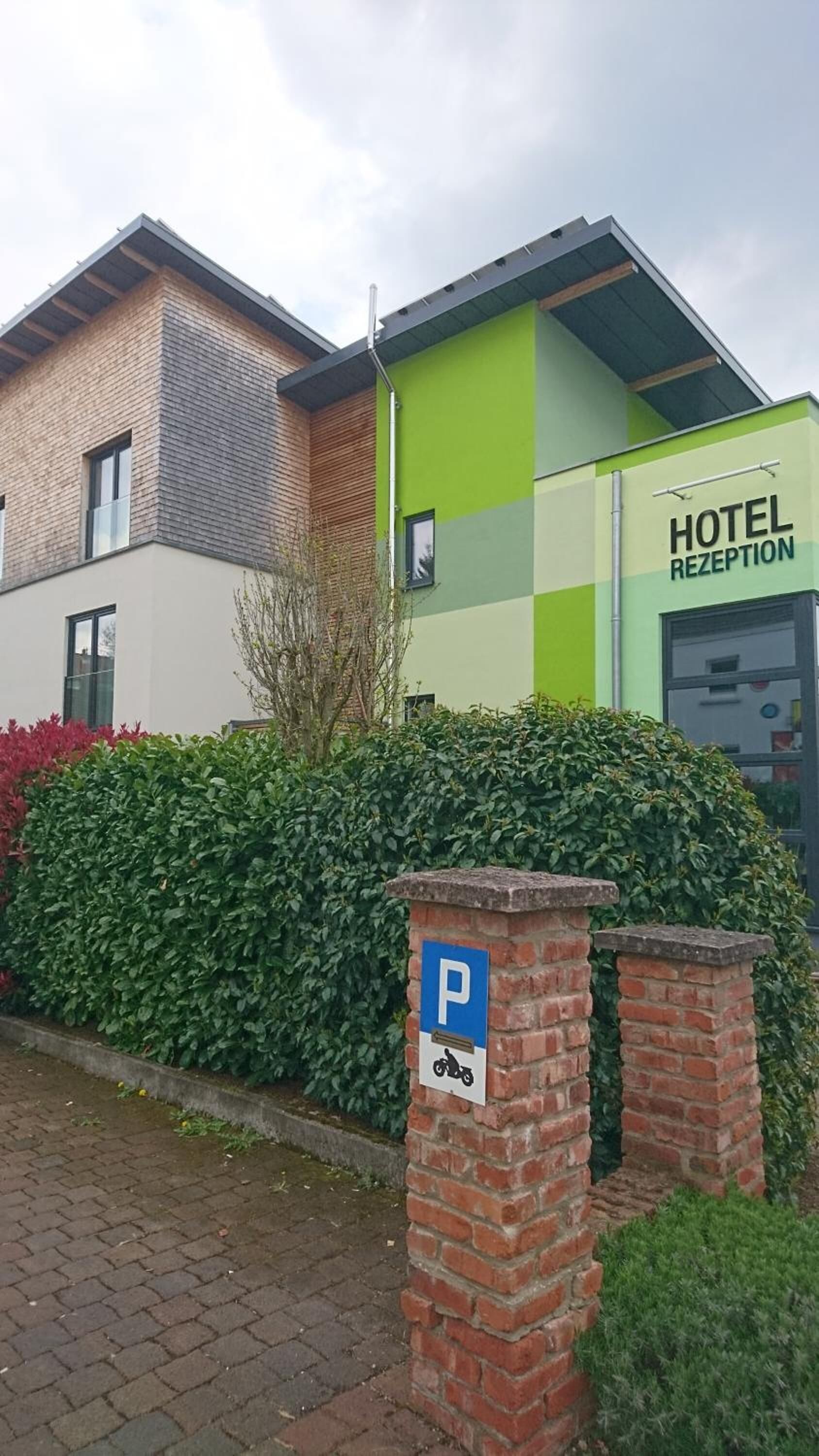 A hotel reception with a modern design, featuring a green and white facade, and a parking sign indicating a parking area for motorcycles.
