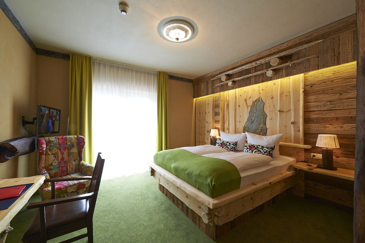 A wooden bed with a green blanket, a wooden nightstand with a lamp, a chair with a colorful cushion, and a painting on the wall.