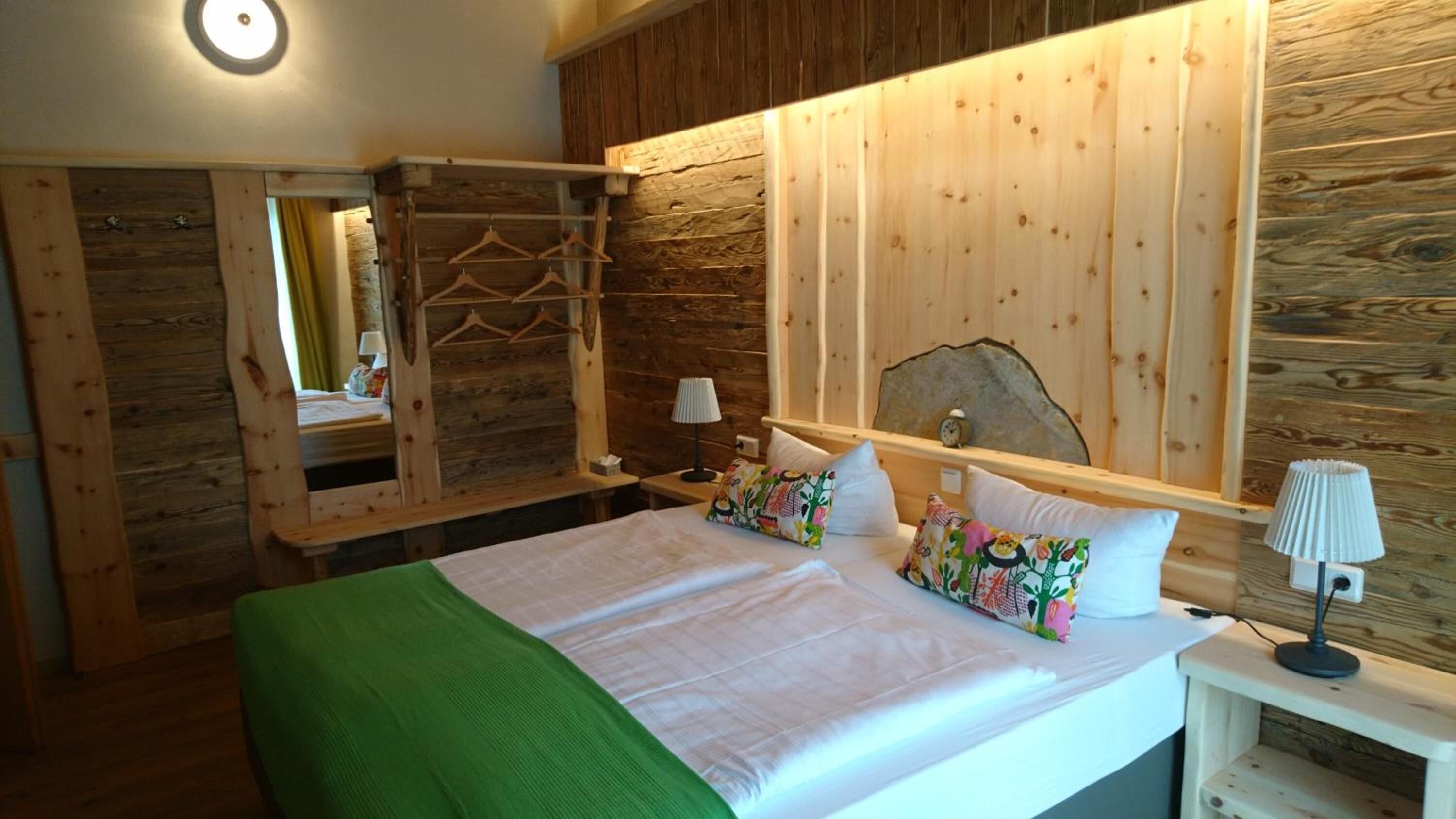 Una habitación de madera con una cama, un espejo, un perchero y una mesita de noche con una lámpara.