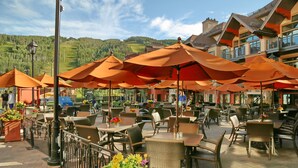 Un café al aire libre con mobiliario de mimbre y sombrillas naranjas, con una montaña y un edificio con balcones de fondo.