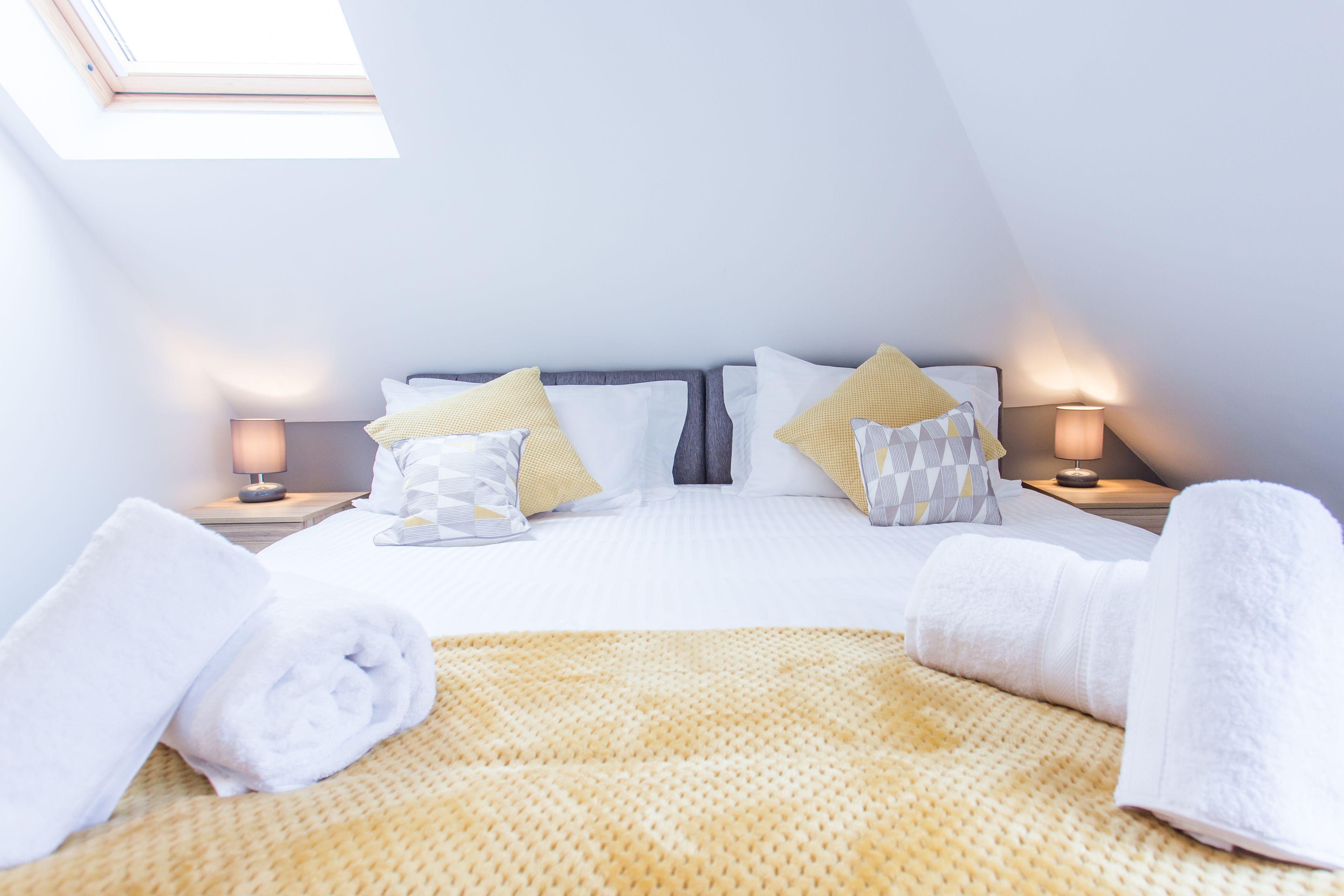 Un lit bien fait, avec du linge de lit blanc et une couverture jaune à motifs, deux lampes de chevet et une lucarne au-dessus.