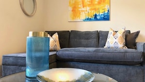 A modern living room with a grey sectional sofa, a glass coffee table, a blue vase, and a painting on the wall.