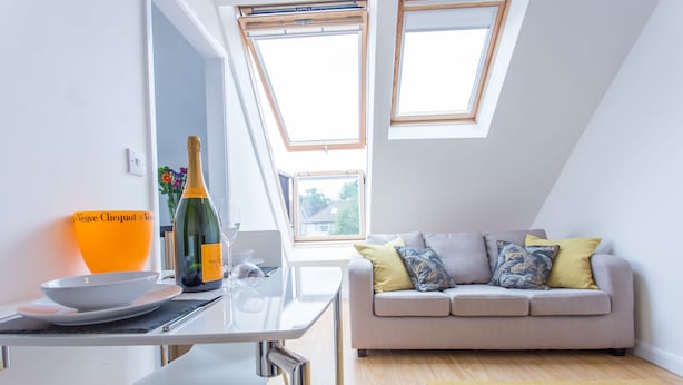 A modern living room with a grey sofa, a dining table with a bottle of Veuve Clicquot, and a yellow bowl.