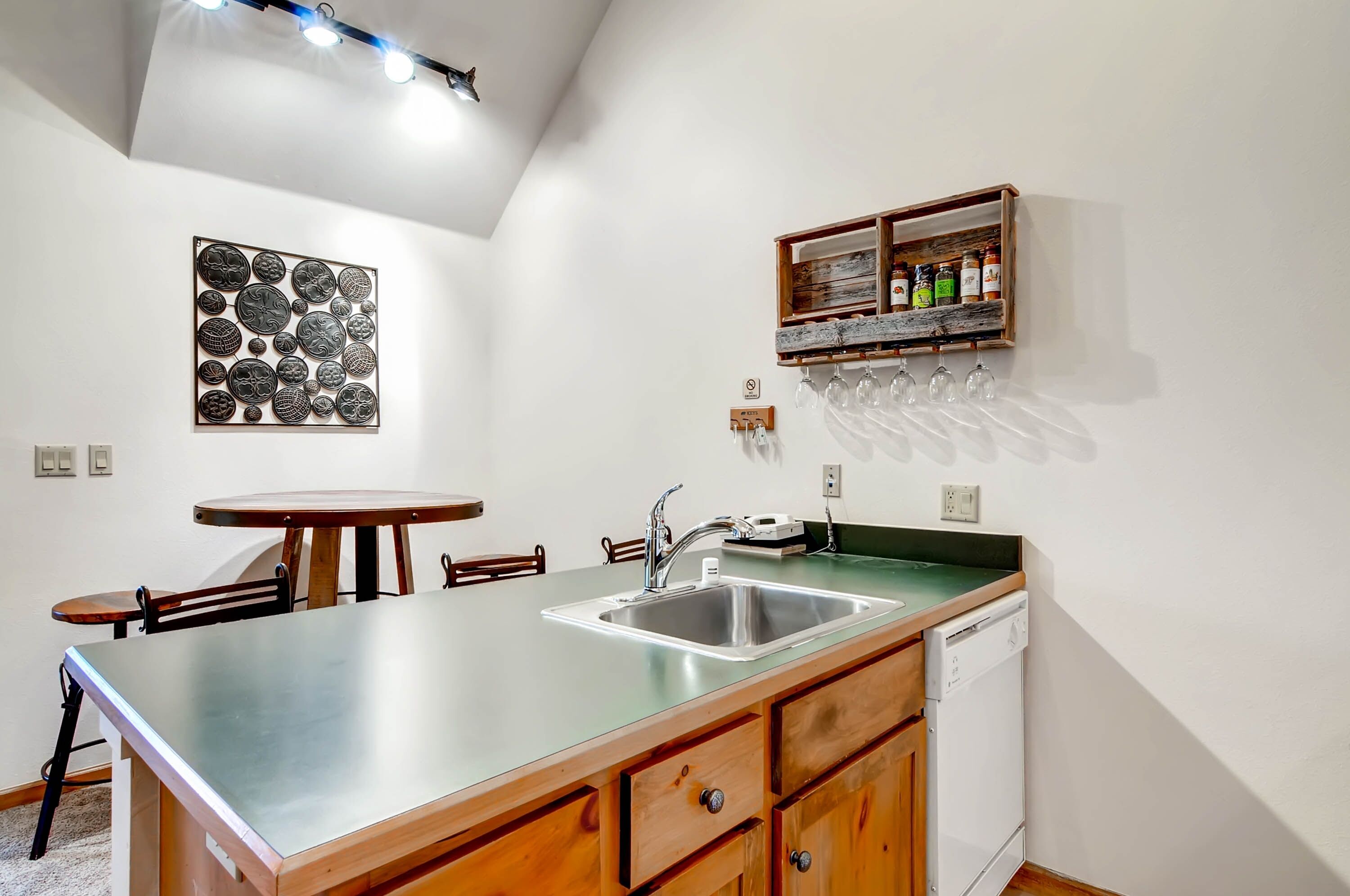 Una cocina con un lavamanos de acero inoxidable, armarios de madera, una mesa redonda para comer y una estantería para vinos montada en la pared.