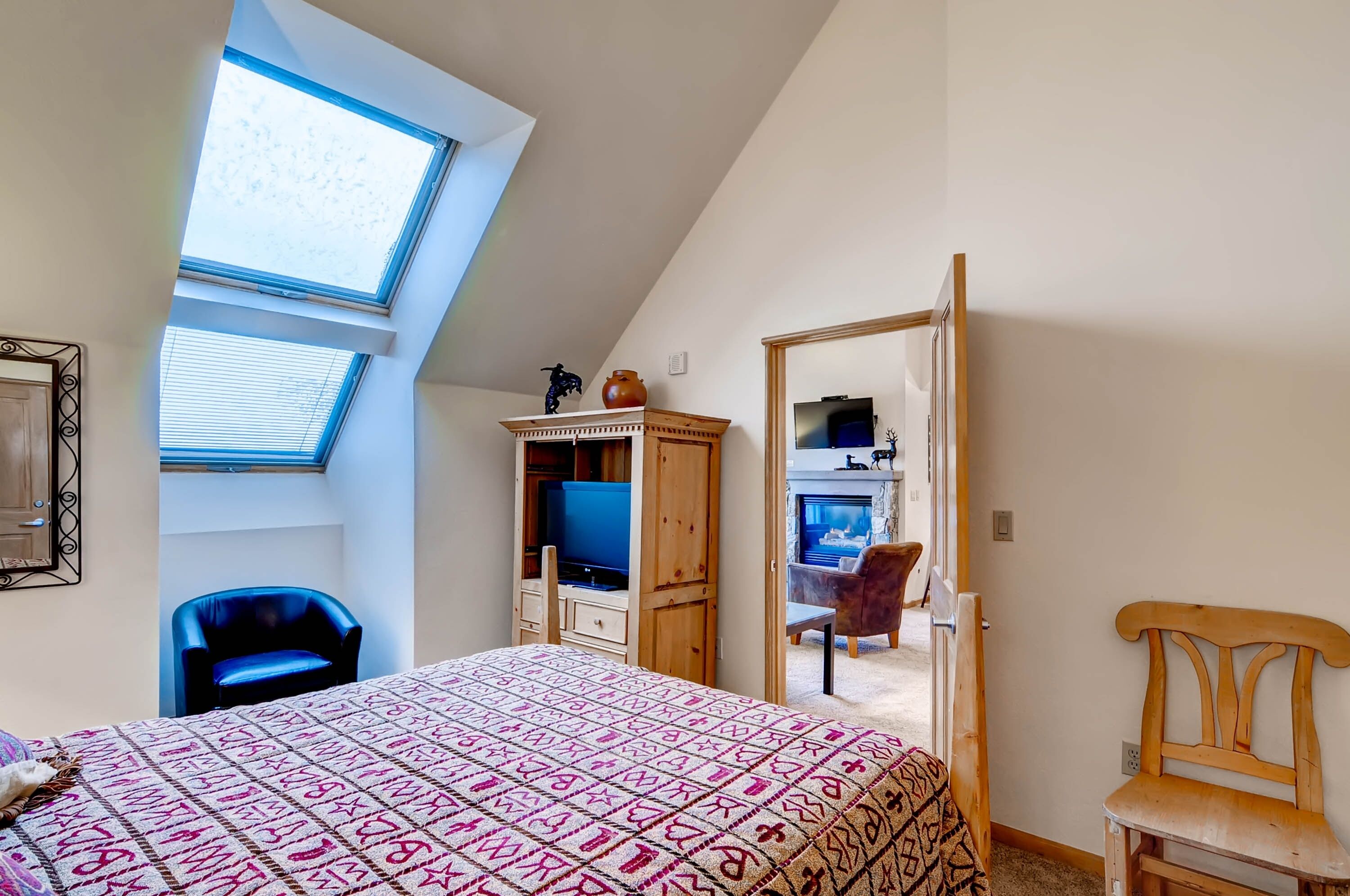Une chambre avec un plafond en pente, un lit recouvert d’une couvre-lit à motifs, un téléviseur, une armoire en bois et une chaise.