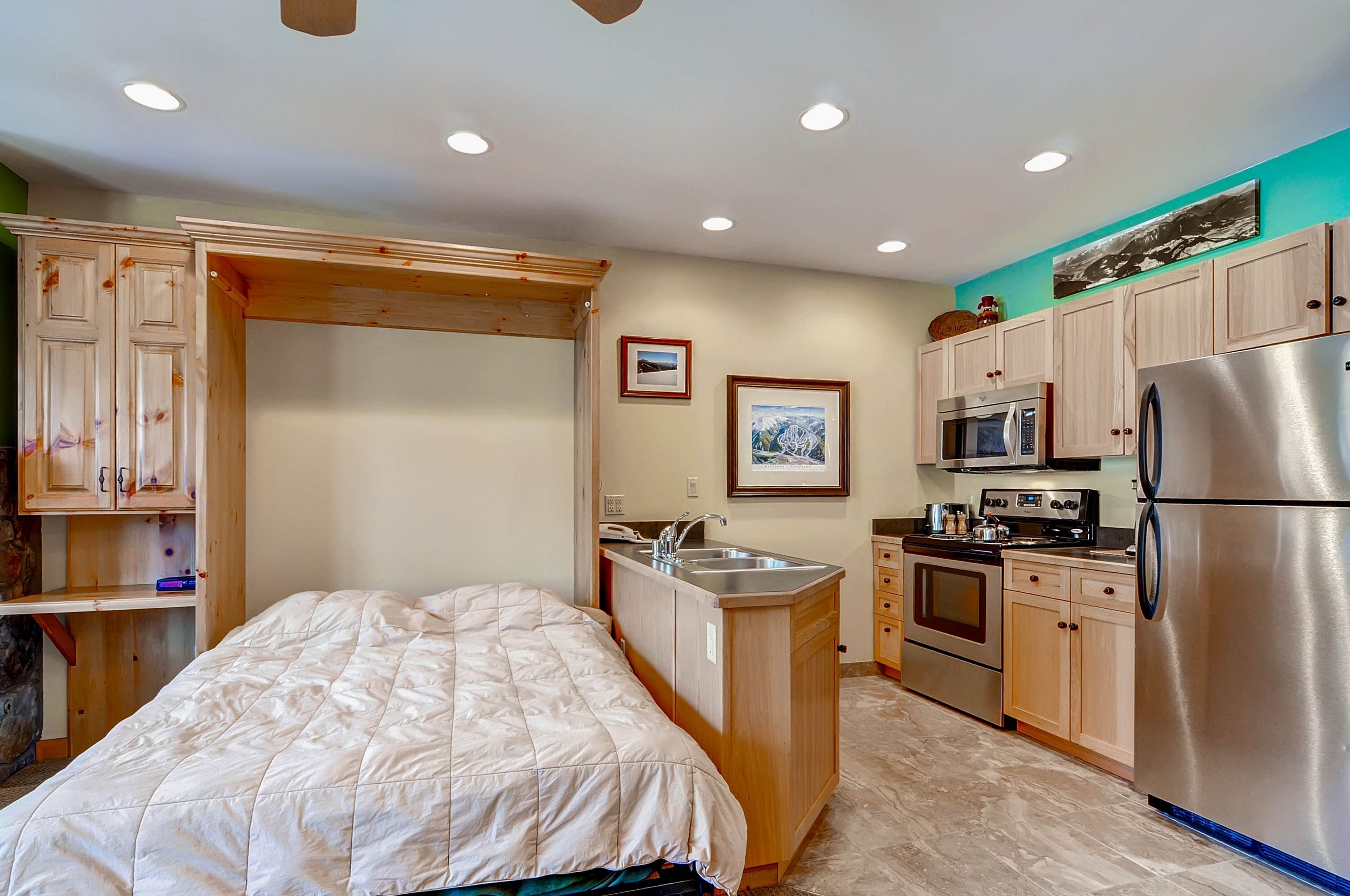 A compact kitchen with a bed, wooden cabinets, a stainless steel refrigerator, and a microwave.