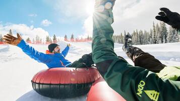 A snowy landscape with a person in a blue jacket and another in a green jacket riding a red snow tube.