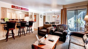 A modern living room with a kitchen area, featuring dark wooden furniture, a coffee table, and a large window with curtains. - Spacious, Upgraded 2 Bdr W/ Fireplace & Hot Tub Access 2 Bedroom Condo by RedAwning (Keystone)