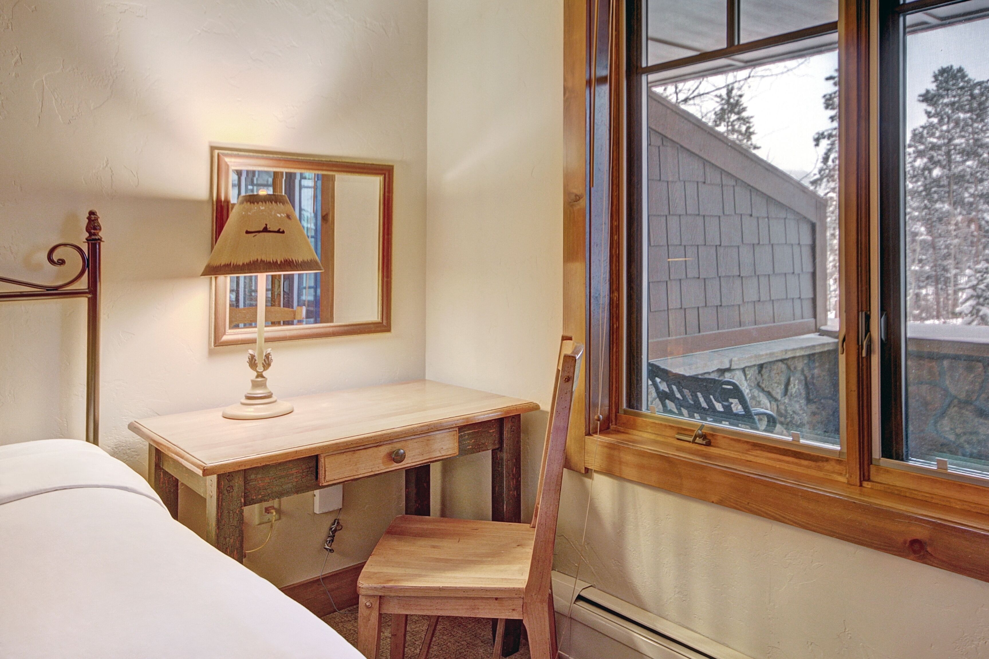Ein Schlafzimmer mit einem Holztisch, einem Stuhl, einer Lampe und einem Fenster mit Blick auf ein Backsteinhaus.