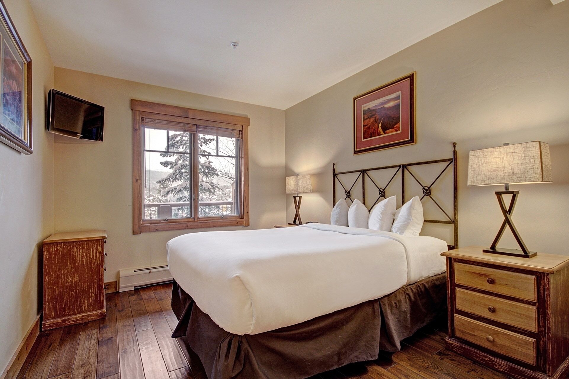 Une chambre à coucher avec un grand lit, une table de chevet en bois, une lampe de chevet, un tableau encadré au mur et une fenêtre donnant sur des arbres.