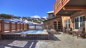 Un área de piscina con baldosas de piedra, terraza de madera y vistas a montañas nevadas.