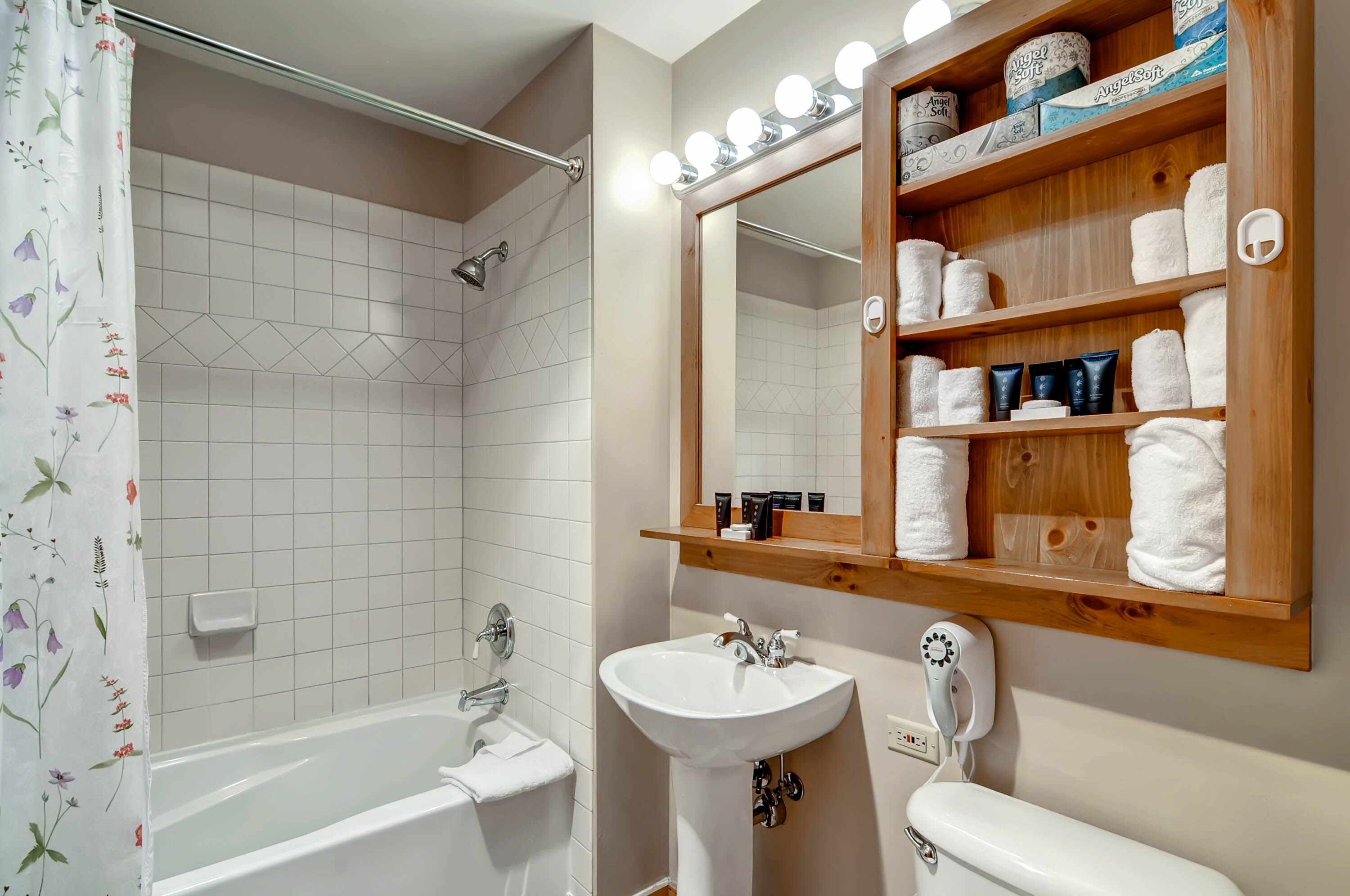 A bathroom with a bathtub, a sink, a toilet, a shower, and a vanity with a mirror and shelves of toiletries.
