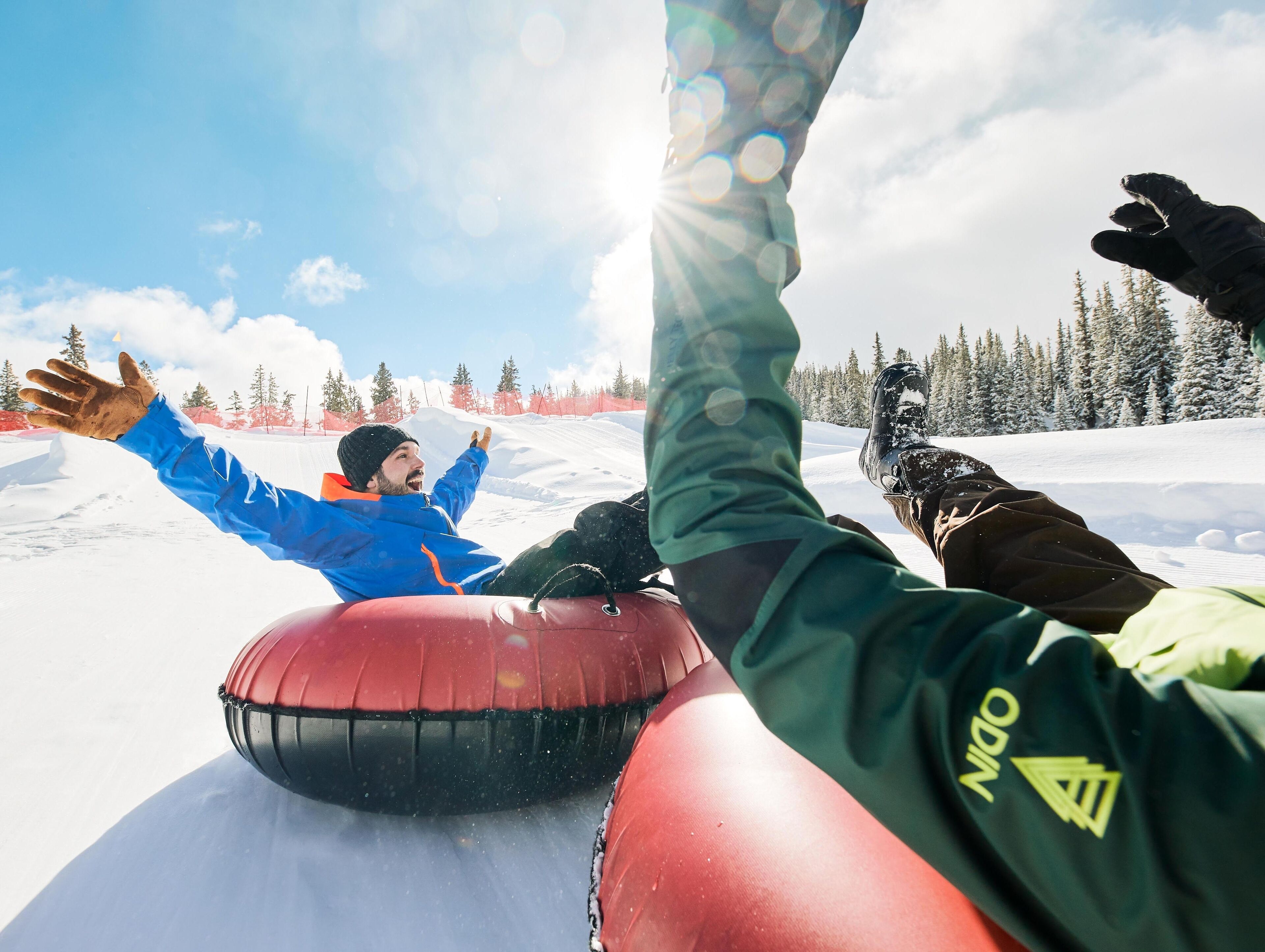 A snowy landscape with a person in a blue jacket and another in a green jacket riding a red snow tube.