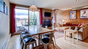 A modern living room with a wooden floor, a dining area with a wooden table and chairs, a fireplace, a sofa, and a television mounted on the wall. - Plaza 1407 (Keystone)