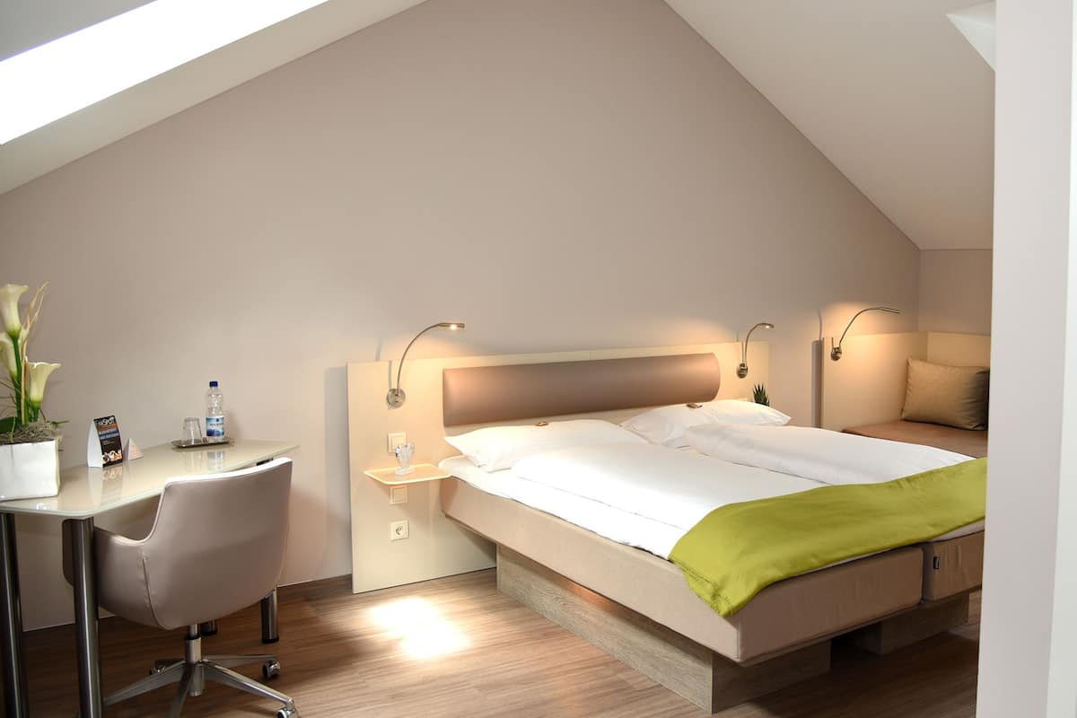 A modern hotel room with a sloped ceiling, a bed with a green throw, a desk with a chair, and bedside lamps.