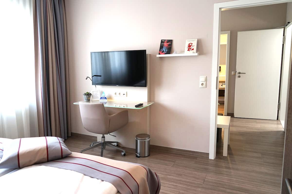 A hotel room with a bed, a desk with a computer, a TV, and a small shelf with books.