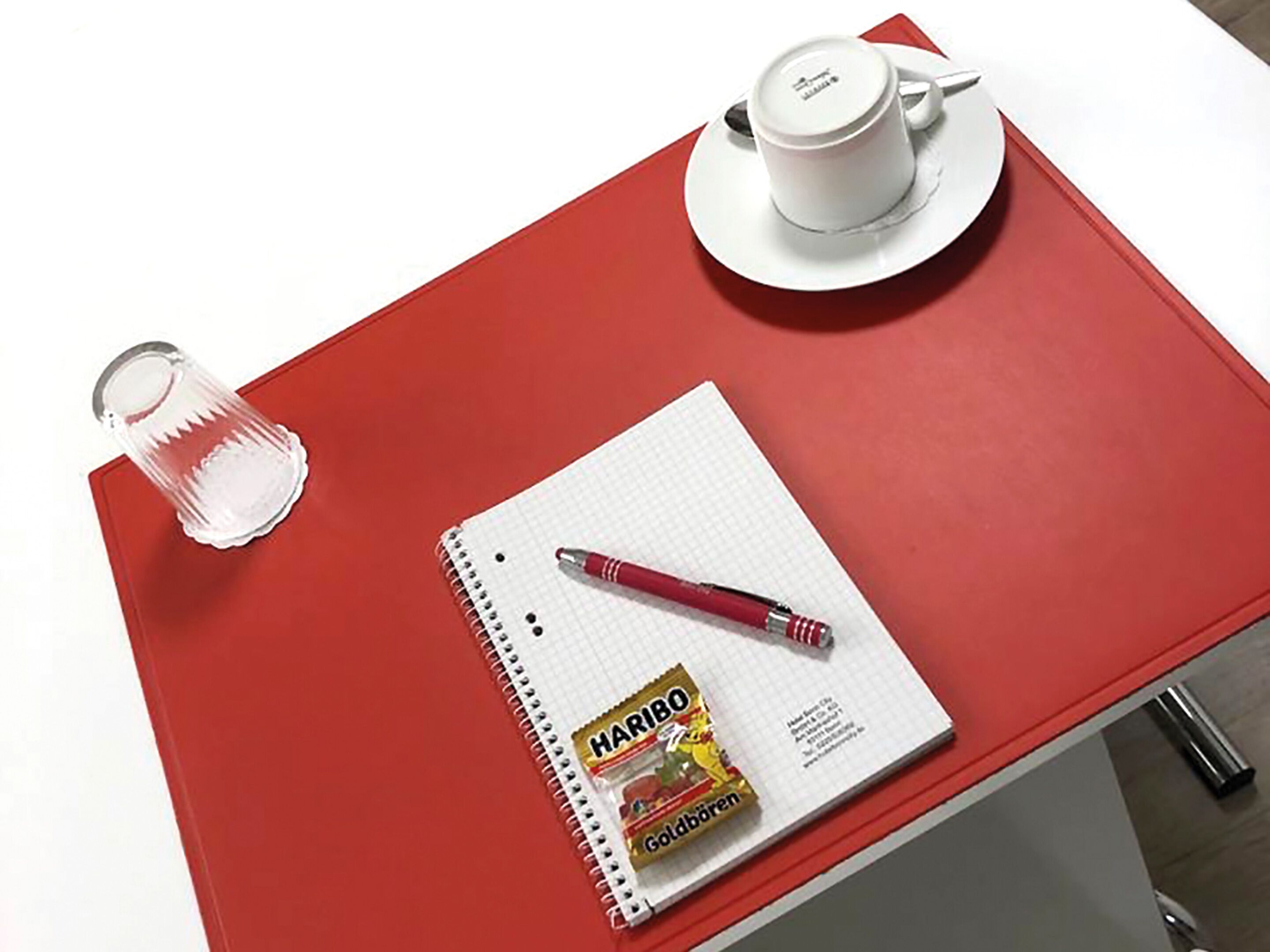 Een rode tafel met een wit notitieboek, een pen, een glas water en een witte kop met schotel.