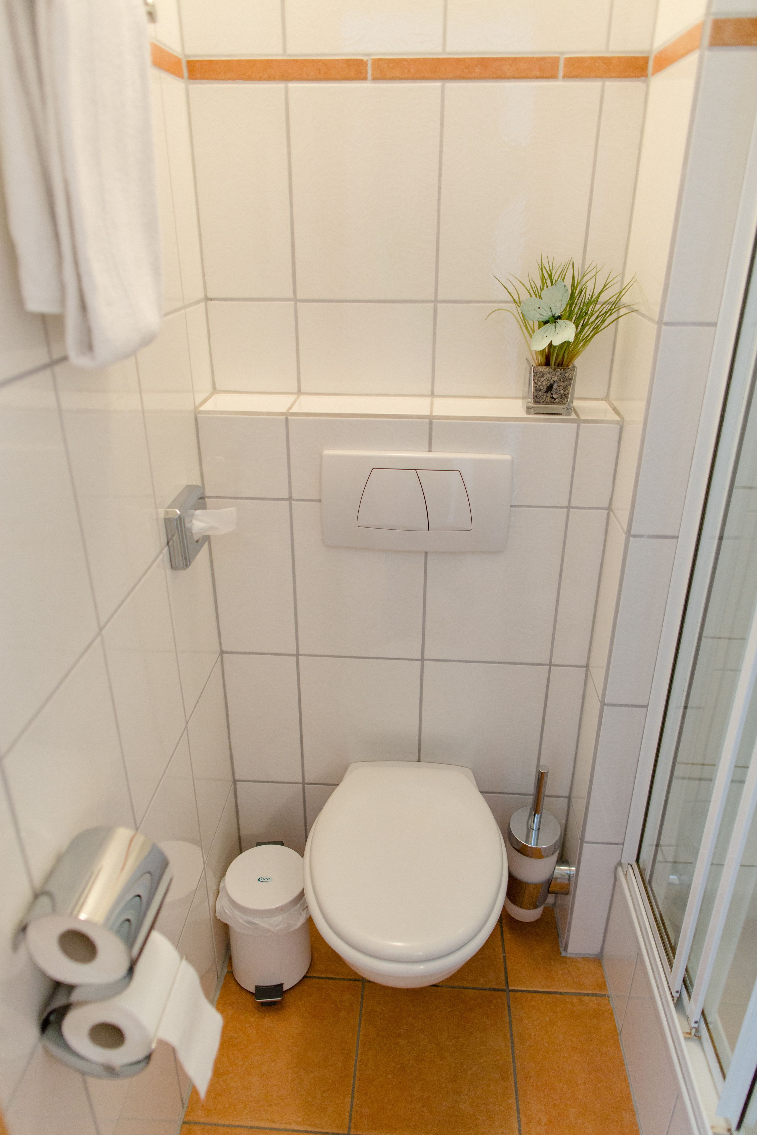 Ein kleines Badezimmer mit Toilette, Mülleimer, Toilettenpapierhalter und einer kleinen Pflanze an der Wand.