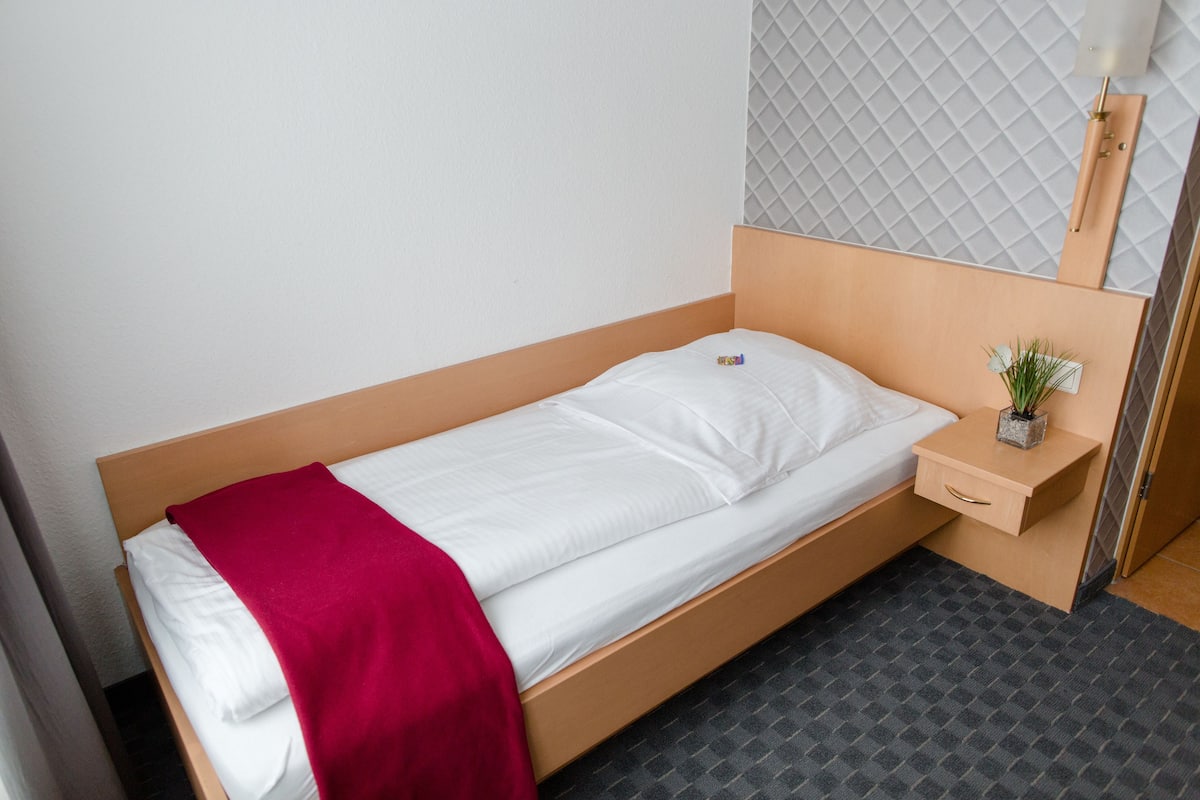 A single bed with a wooden frame, a white mattress, and a red blanket. There is a small bedside table with a plant and a drawer. The room has a grey patterned wall and a grey floor.