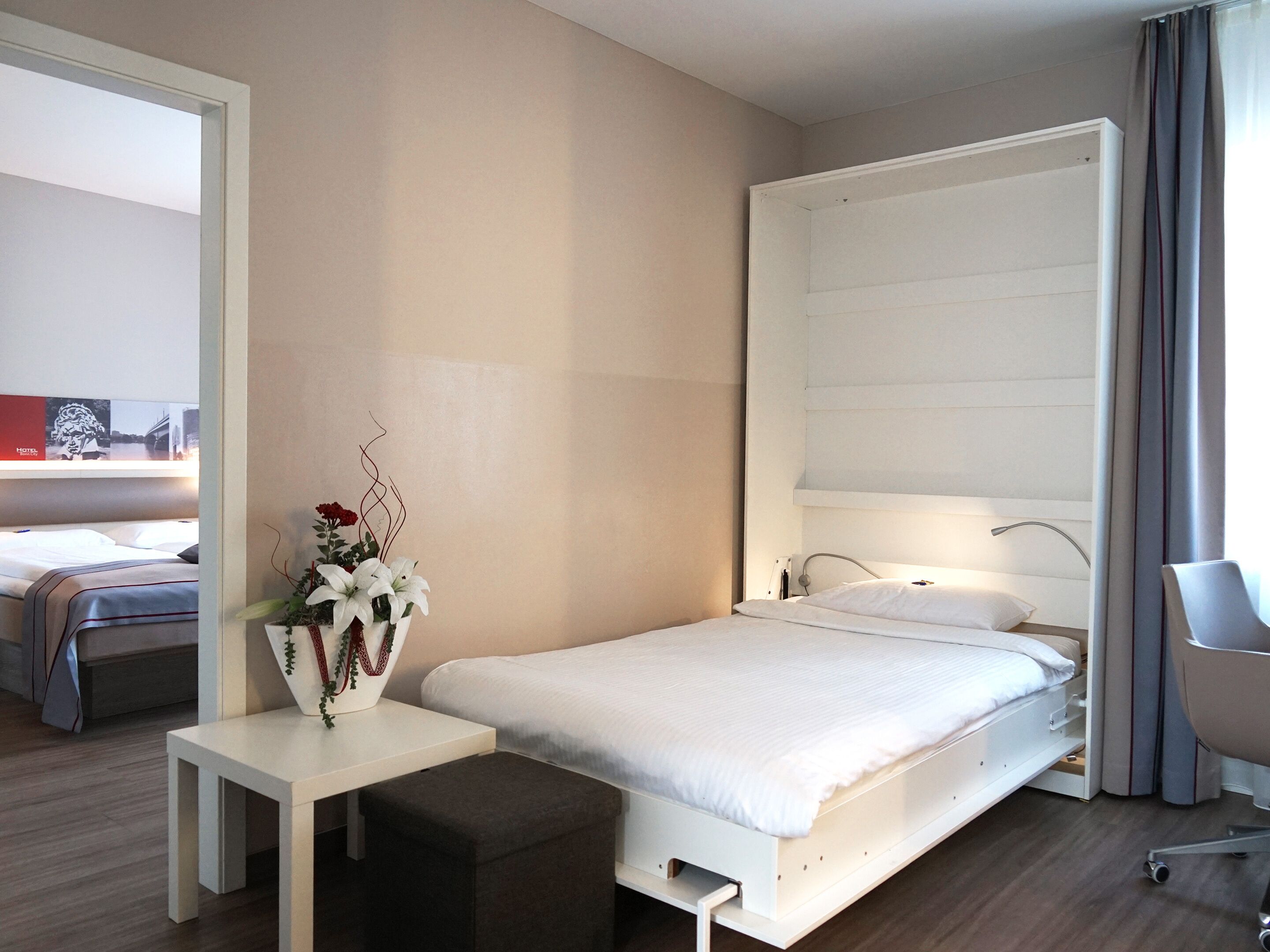 Habitación de hotel moderna con una cama individual, una mesita de noche pequeña, una silla y un ventanal con cortinas.