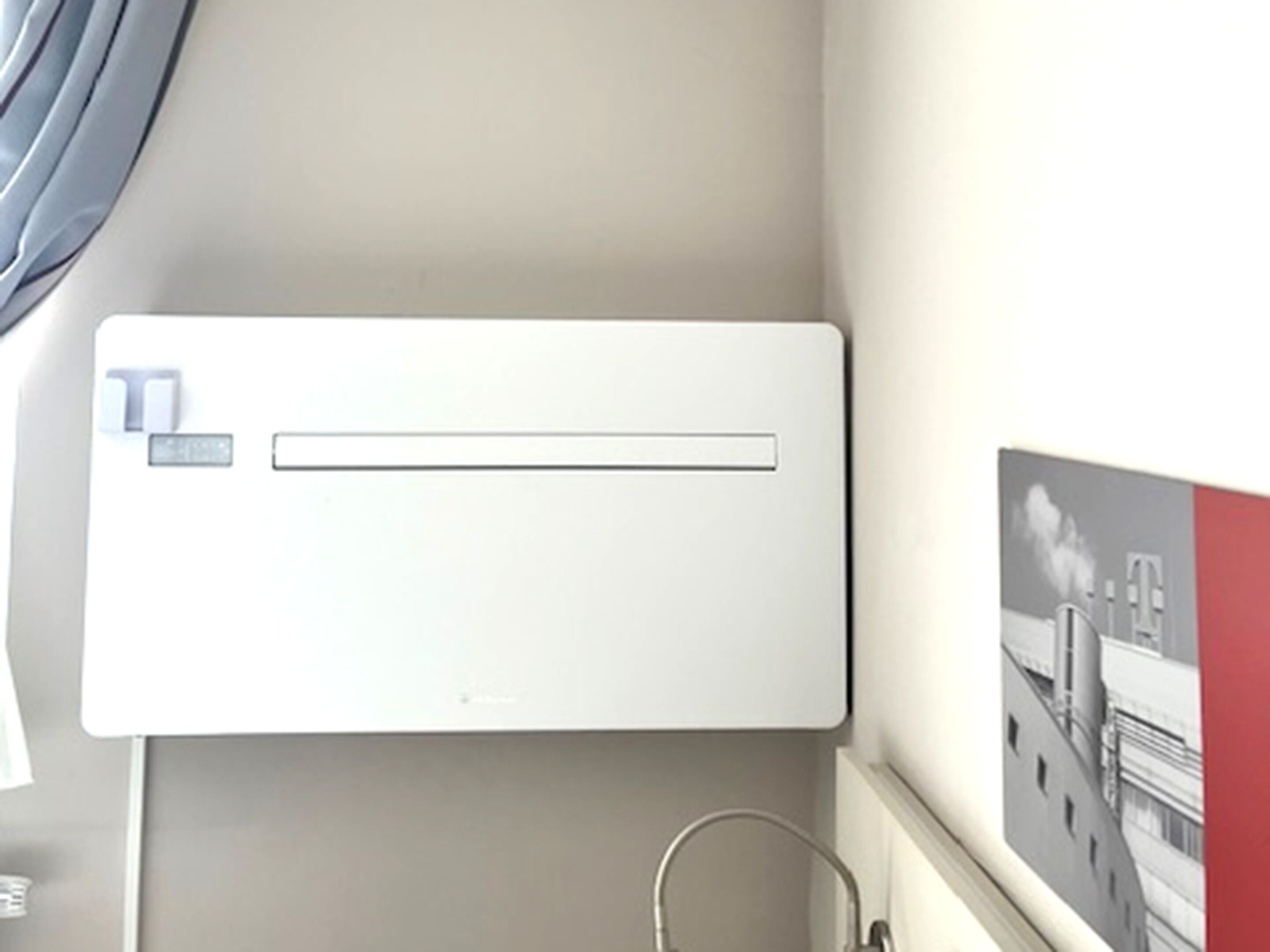 Een witte airconditioningunit aan een muur, met een raam en een fotolijst.