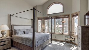 Een hemelbed met een geruit sprei, een nachtkastje met een lamp en een stoel bij een raam met uitzicht op bomen.
