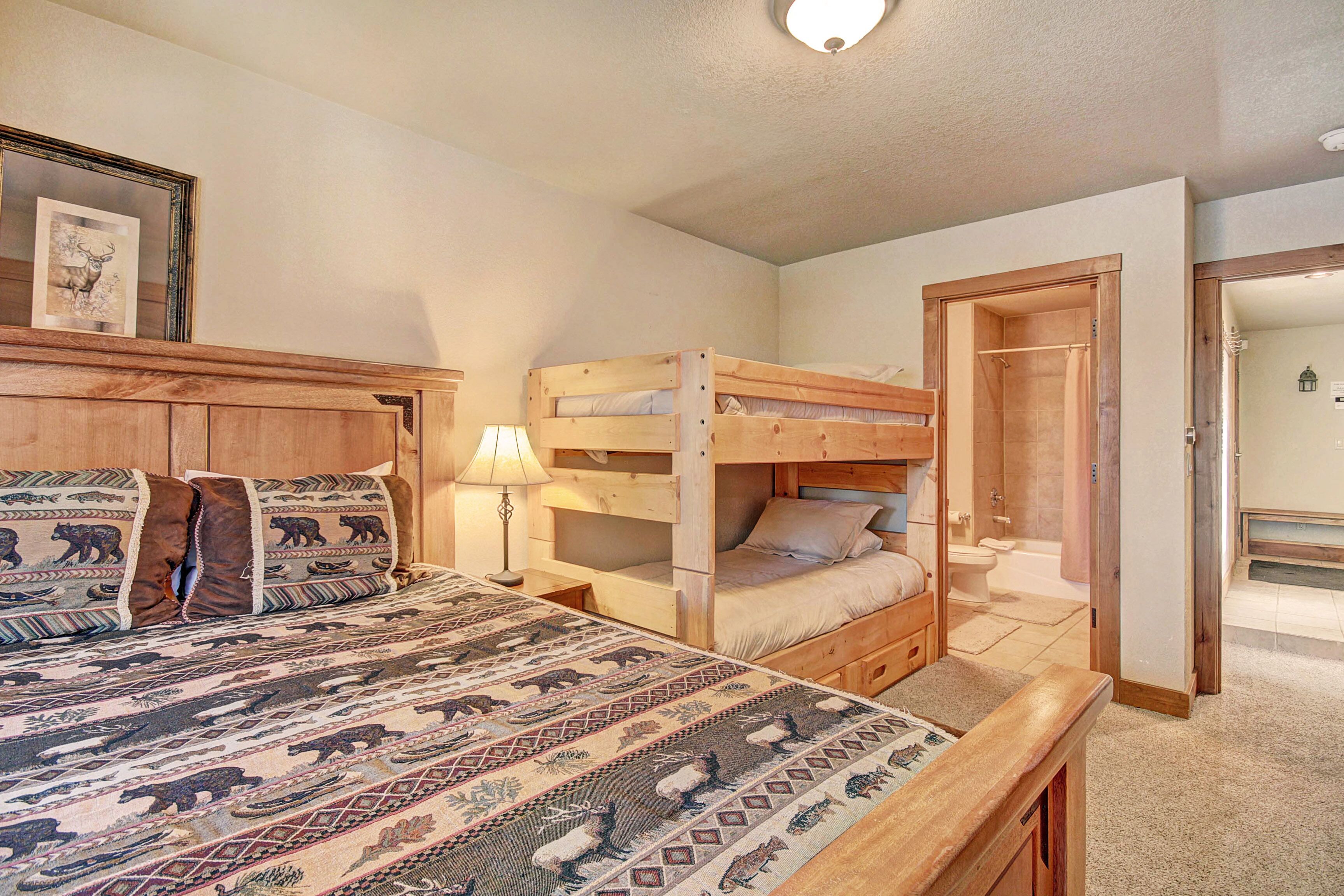 A bedroom with a large bed, a smaller bunk bed, a wooden headboard, a bedside table with a lamp, and a door leading to a bathroom.