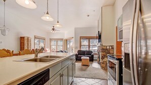 A kitchen with a dishwasher, double sink, and a stone fireplace. There is a dining area with wooden chairs and a sofa.