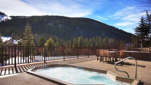 A hot tub with a view of a forested mountain, a wooden deck, and outdoor furniture.