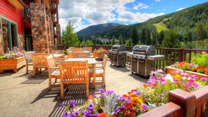 An outdoor patio with wooden furniture and barbecue grills, surrounded by colorful flowers and a mountainous backdrop.