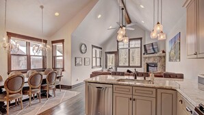 A spacious kitchen with a large island, wooden dining table, and chairs. There is a stone fireplace, a ceiling fan, and pendant lights.
