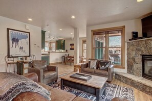 A spacious living room with a fireplace, a kitchen in the background, and a large poster on the wall. - 201 Mill Creek (72 River Run Road) 4 Bedroom Condo by RedAwning (Keystone)
