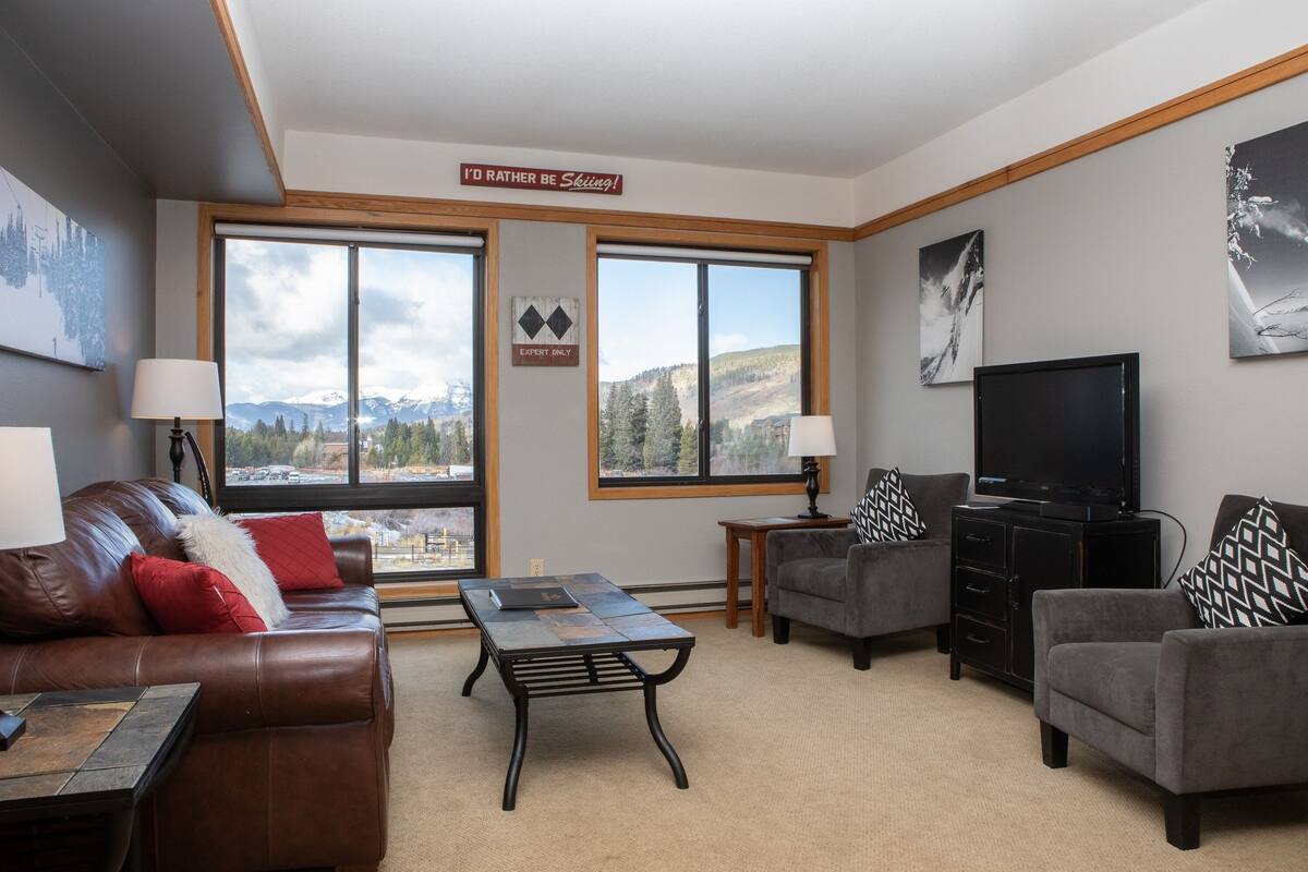 A living room with a brown leather sofa, a grey armchair, a coffee table, and a television. There are two windows with a view of mountains and a sign that says "I'd rather be skiing!"